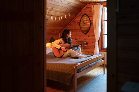 Foto de Pensive lonely woman sits on large cozy bed with guitar in hands, plays melody, sadly looks out window, singing favourite song. Rest in country wooden house. Female enjoying playing musical instrument - Imagen libre de derechos