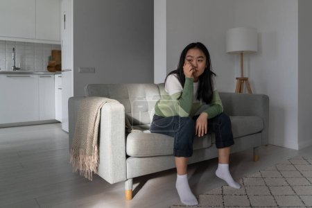 Foto de Upset unhappy Asian woman sits on couch propping head with hand and looking into distance without initiative. Unmotivated sad Korean girl suffering from depression spending time alone in living room - Imagen libre de derechos