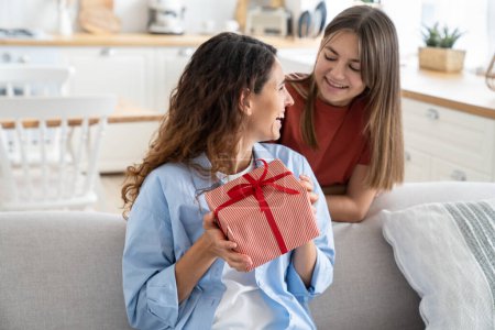 Photo for Cute teen girl loving daughter congratulating happy young mother with birthday at home, give her gift box with red ribbon. Child surprising mom on Mothers Day, kid making mum happy. Family holidays - Royalty Free Image