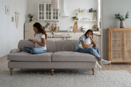 Foto de Focused busy woman mother using smartphone during family time child daughter at home. Working from home mom looking and mobile phone screen, ignoring sad upset teen girl kid. Parents and screen time - Imagen libre de derechos