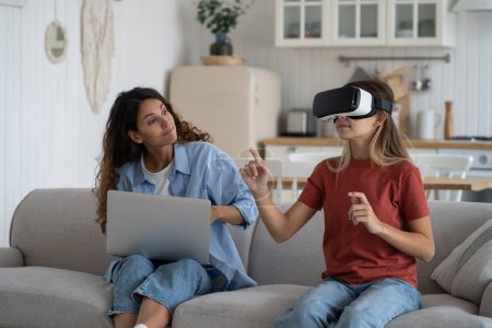Téléchargez les photos : Mother asking daughter in VR headset to take game play break while sitting together on sofa at home, child spending long time immersed in virtual world. Health dangers of virtual reality for children - en image libre de droit