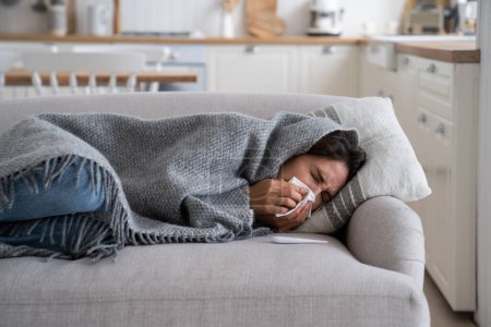 Téléchargez les photos : Young woman lying under blanket on sofa coughing and sneezing, using paper tissue to blow her nose. Unhealthy female caught cold or flu, resting on couch, suffering coronavirus. Cold homes and health - en image libre de droit