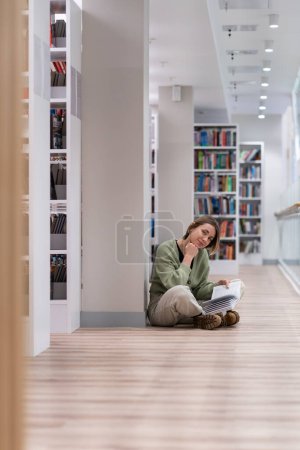 Photo for Positive inquisitive mature woman looking at camera sits on floor in library enjoying reading fiction. Optimistic casual girl posing in bookstore near shelves with textbooks, leaning chin on hand - Royalty Free Image