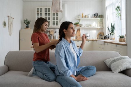 Foto de Teen daughter spending time with mom at home, braiding mothers hair while relaxing together on sofa. Young happy woman mother bonding with teenage girl, enjoying motherhood. Mother-daughter friendship - Imagen libre de derechos