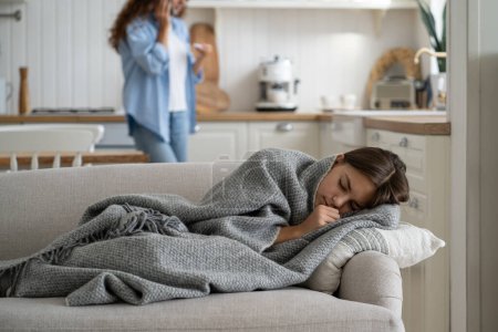 Photo for Sick repressed teen girl sleeping in house on couch wrapped in blanket warming up in cold weather. Caring woman calling by phone holding thermometer standing behind schoolgirl lying with closed eyes - Royalty Free Image