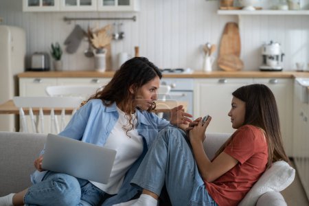 Photo for Confused teenage girl looking at mother scolding for using mobile phone too long. Caring attentive woman with laptop on knees sits on sofa of own apartment next to daughter with smartphone. Mistrust. - Royalty Free Image