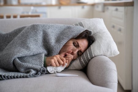 Foto de Young woman lying under blanket on sofa coughing and sneezing, using paper tissue to blow her nose. Unhealthy female caught cold or flu, resting on couch, suffering coronavirus. Cold homes and health - Imagen libre de derechos