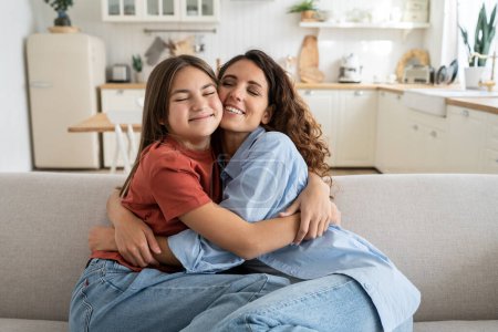 Téléchargez les photos : Happiness in motherhood. Joyful young mother and teen daughter cuddling hugging while sitting on sofa together, excited overjoyed girl child embracing mom coming back home. Family wellbeing concept - en image libre de droit