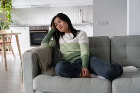 Foto de Relajada mujer asiática soñadora sonriendo y mirando hacia otro lado mientras se sienta en el sofá en casa, mujer despreocupada descansando en el sofá, sintiéndose tranquila y feliz, tomando un descanso de todo. Concepto de bienestar mental - Imagen libre de derechos