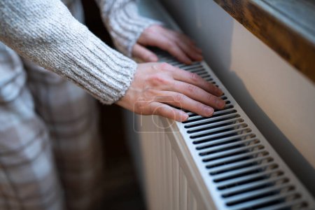Foto de Closeup of woman in woolen sweater wants to keep warmth in apartment. Female puts hands on room central heating battery to warm up and prevent illness because of coldness. Person checks work of heater - Imagen libre de derechos