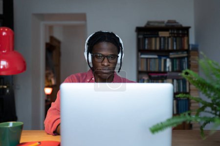 Téléchargez les photos : Focused black man freelancer looking at laptop screen working in evening at home office, unhappy tired young African student guy feeling overwhelmed with online classes. Freelance and remote learning - en image libre de droit