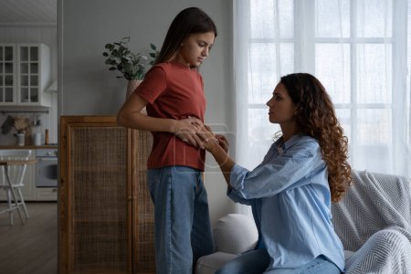 Téléchargez les photos : Sickly young school-age girl approaches mother to complain of abdominal pain or problems with digestive system. Restrained casual Woman Helps Daughter Cope with Teenage Health Issues located at home - en image libre de droit