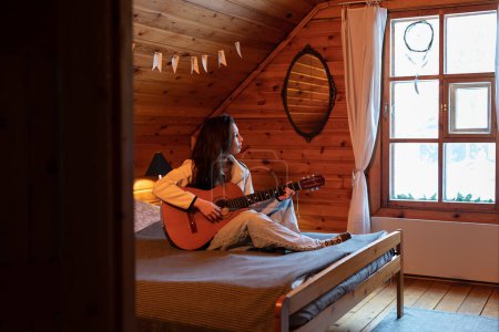 Photo for Creative millennial girl sitting on bed holding guitar in hands and looking out of window, waiting for inspiration. Musical Instrument and mental health. Hobby and recreational activity concept - Royalty Free Image