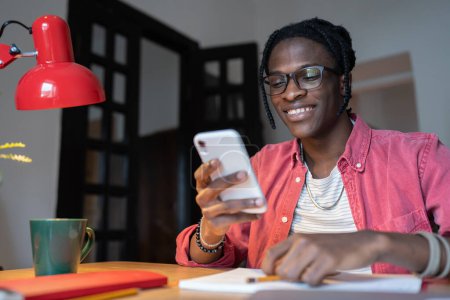 Glücklich optimistischer afroamerikanischer Mann smm Spezialist verwendet Mobiltelefon, um soziale Medien des Unternehmens zu verwalten. Selbstgenügsam lächelnder Black-Guy-Vermarkter sitzt am Bürotisch und wirbt mit Blog für Unternehmen