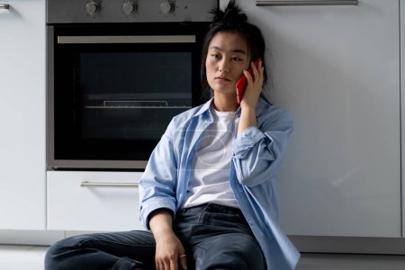 Téléchargez les photos : Depressed young Asian woman sitting on floor at home constantly calling ex-boyfriend after breakup, feeling lonely, talking with close friend over phone getting support while going through divorce - en image libre de droit