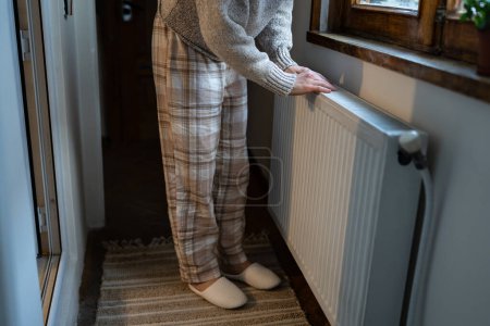 Foto de Closeup of woman warming her hands on the heater at home during cold winter days. Female getting warm up her arms over radiator. Concept of heating season, cold weather. - Imagen libre de derechos