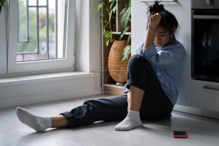 Unglücklich deprimiertes asiatisches Mädchen sitzt zu Hause auf dem Boden und schaut auf das Smartphone, das auf einen Anruf vom Freund wartet, fühlt sich traurig und herzzerreißend, nachdem sie eine SMS erhalten hat, kann Ex nicht vergessen