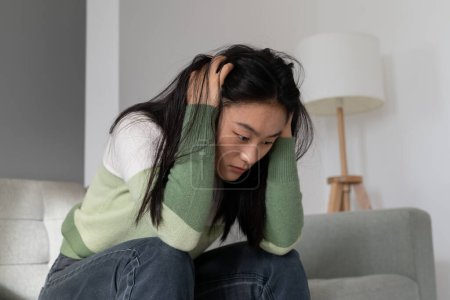 Téléchargez les photos : Anxious Asian millennial girl sitting on sofa at home holding head in hands, dealing with depression after abortion, worrying about making wrong decision. Unexpected pregnancy concept - en image libre de droit