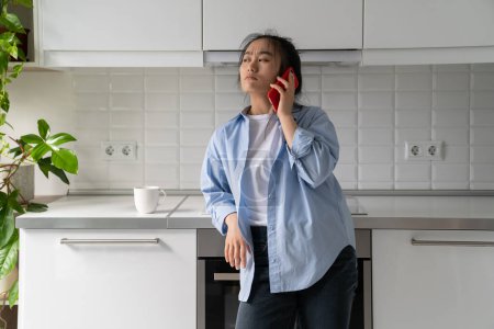 Foto de Preocupado confundido milenial chica asiática teniendo una conversación desagradable en el teléfono móvil, mujer coreana insatisfecha ama de casa sosteniendo teléfono inteligente esperando en espera mientras está de pie en la cocina en casa - Imagen libre de derechos