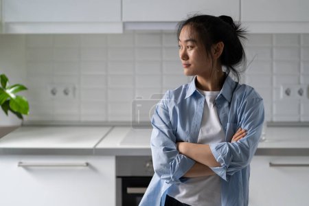 Photo for Relaxed dreamy Asian woman smiling and looking away thinking about future plans standing in kitchen. Optimistic young korean girl posing with arms crossed remembering happy moments from vacation - Royalty Free Image