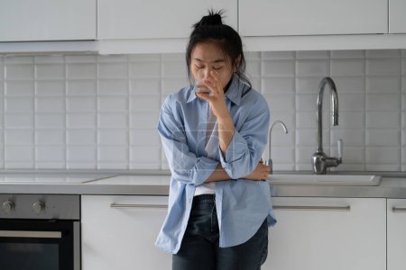Téléchargez les photos : Stressed nervous young Asian woman standing in kitchen drinking water calming down after panic attack at home. Korean millennial girl feeling thirsty. Dehydration and anxiety - en image libre de droit