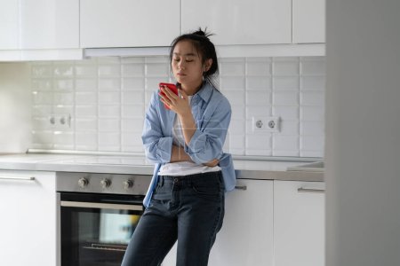 Téléchargez les photos : Bouleversé jeune femme asiatique tenant smartphone devient émotionnellement triste en lisant des nouvelles déprimantes sur Internet, debout dans la cuisine, inquiet femme regardant l'écran mobile recherche d'informations en ligne - en image libre de droit