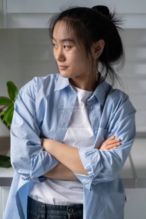 Photo for Serious confident young Asian girl standing at home with arms crossed looking away thinking of future, doing visualization techniques. Pensive millennial woman housewife pondering, lost in thoughts - Royalty Free Image