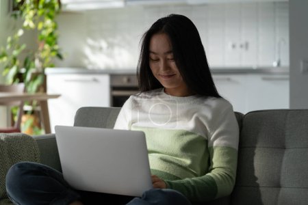 Photo for Smiling delighted millennial Asian girl sitting on sofa at home looking at laptop screen, shopping online. Happy young woman having conversation on dating website, typing on keyboard, selective focus - Royalty Free Image