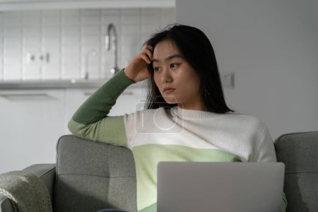 Foto de Worried anxious Asian girl sitting with laptop on sofa looking aside, sad female freelance worker feeling lonely and depressed while working remotely from home, unproductive Korean woman freelancer - Imagen libre de derechos