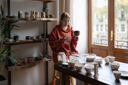 Photo for Young woman potter resting in cozy home pottery studio, drinking tea and using smartphone. Female ceramist sitting at work table, taking break. Ceramic teacher having coffee break between lessons - Royalty Free Image