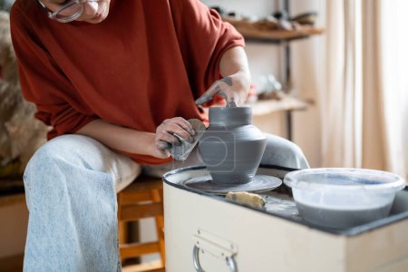 Photo for Attentive woman sits in pottery workshop and diligently does what she loves, which brings income and pleasure. Female makes aesthetic items for home with own hands. Working with clay for mental health - Royalty Free Image