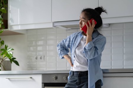 Téléchargez les photos : Worried upset Asian woman housewife standing alone in kitchen making unpleasant phone call, talking on smartphone with concerned face expression. Sad mother calling child who ignoring parent call - en image libre de droit