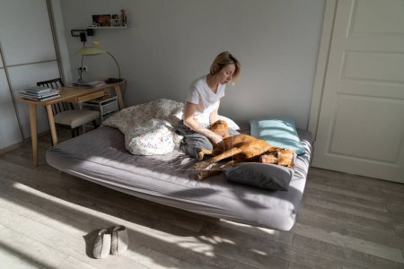 Téléchargez les photos : Melancholy pensive woman sits on bed and petting dog to get rid of depression after getting psychological trauma. Lonely mature lady with blond hair plays with beloved pet in bedroom of own apartment - en image libre de droit