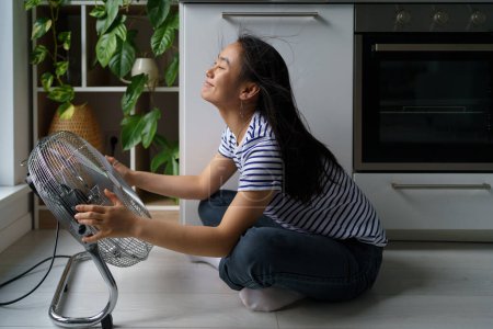 Photo for Cheerful Asian girl enjoys cold wind from electric fan sits on floor in kitchen. Young Japanese woman resting at home and sits by fan and enjoys the cool breeze. Summertime, heat concept. - Royalty Free Image