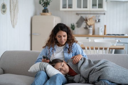Téléchargez les photos : Loving caring mother woman sits on sofa in living room and smiles looking at teenager daughter falling asleep on knees. Happy kind casual girl nanny admires sleeping girl covered with warm blanket - en image libre de droit