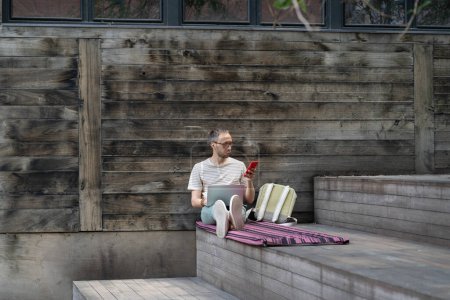 Photo for Young enterprising man programmer sits outdoors with laptop and phone creating software to sell online. Successful guy IT engineer located near wooden wall of building on street in summer weather - Royalty Free Image