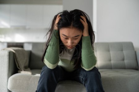 Photo for Unhappy Asian woman sits on sofa at home feeling sad, frustrated and lonely, dealing with breakup or divorce. Depressed Korean girl thinking of relationship trouble. Mental health, depression, anxiety - Royalty Free Image
