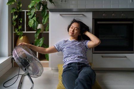 Foto de Smiling Asian woman sits on floor and enjoying air currents coming from electric fan. Carefree Korean girl in striped t-shirt touch hair spending time alone in kitchen in hot apartment. Summertime. - Imagen libre de derechos