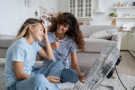 Foto de Satisfied laughing woman and teenage girl enjoy sits on floor in front of turned on fan on summer sunny day. Carefree happy mom and daughter relax in living room after going for walk in hot weather - Imagen libre de derechos