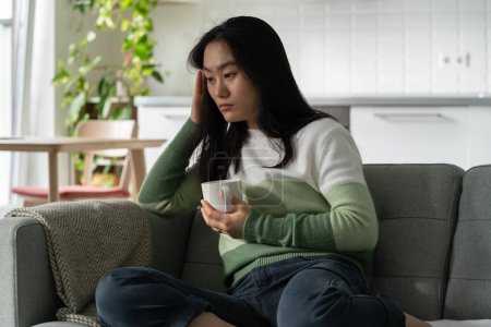 Foto de Deprimido debilitado mujer asiática con taza de café se sienta en el sofá en la sala de estar de su propia casa. triste coreana millennial chica bebe té en la mañana y se siente mal debido a la resaca izquierda después de la noche fiesta - Imagen libre de derechos
