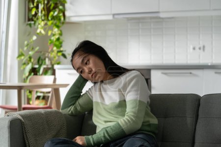 Téléchargez les photos : Depressed suffering Asian woman sits on sofa with head down thinking about failures in personal life. Pessimistic lonely Chinese girl needs to take antidepressants because of fight with boyfriend - en image libre de droit