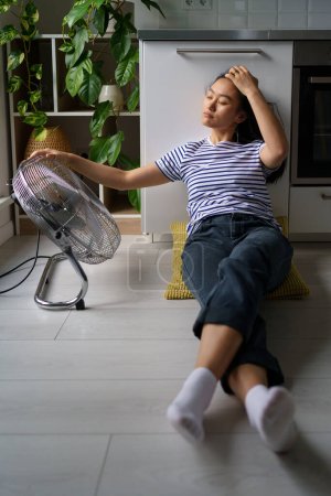 Foto de Relajada joven asiática tumbada en el suelo cerca de ventilador de trabajo y disfruta de frescura y aire fresco. Chica china despreocupada vestida en estilo casual se está enfriando en el día de verano con equipo climático eléctrico - Imagen libre de derechos