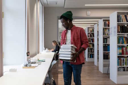 Téléchargez les photos : Libraries and online learning. African American student guy holding pile of books and looking at laptop screen while studying at University library. Learning environment and education concept - en image libre de droit