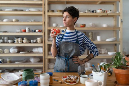 Foto de Creative ceramist woman in dirty apron enjoying her morning coffee from clay mug and looking away while taking break after work in her own cozy pottery studio. Small art business concept - Imagen libre de derechos