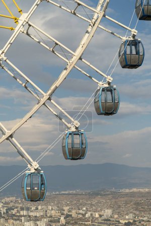 Téléchargez les photos : Ferris wheel with closed cabins fixed on metal frame towering over big city in warm summer weather. Fragment of attraction in amusement park Mtatsminda in capital of Georgia Tbilisi with blue sky - en image libre de droit