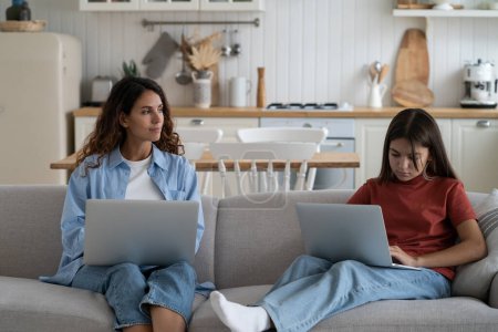 Foto de Young woman mother freelancer working remotely on laptop and supervising remote learning of teenage daughter, balancing work and kids, combining online job and parenting. Family with laptops on sofa - Imagen libre de derechos