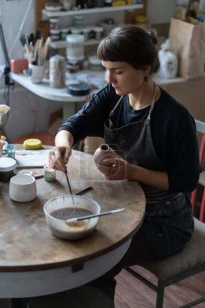 Photo for Concentrated woman ceramist working in workshop sits at table and decorates pottery with colored paints. Professional girl sculptor working on handmade crockery stylized as vintage interior items - Royalty Free Image