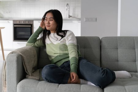 Téléchargez les photos : Carefree satisfied Asian woman enjoying loneliness and silence sits on sofa and remembering happy moments. Charismatic dreamy Japanese girl in casual clothes is waiting for evening to go on date - en image libre de droit