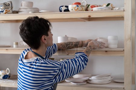 Photo for Enthusiastic pensive woman entrepreneur opens own store with natural clay dishes displaying goods on wall shelves. Successful girl manage small business making pottery and selling to craft shop - Royalty Free Image