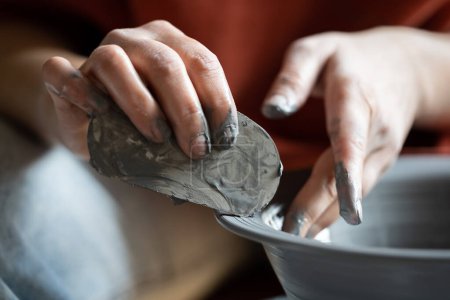 Téléchargez les photos : Hands of crafts woman shaping clay vessel with scraper during production of author dishes for sale in handicraft store. Close-up of fingers of girl owners of pottery workshop stained with clay - en image libre de droit